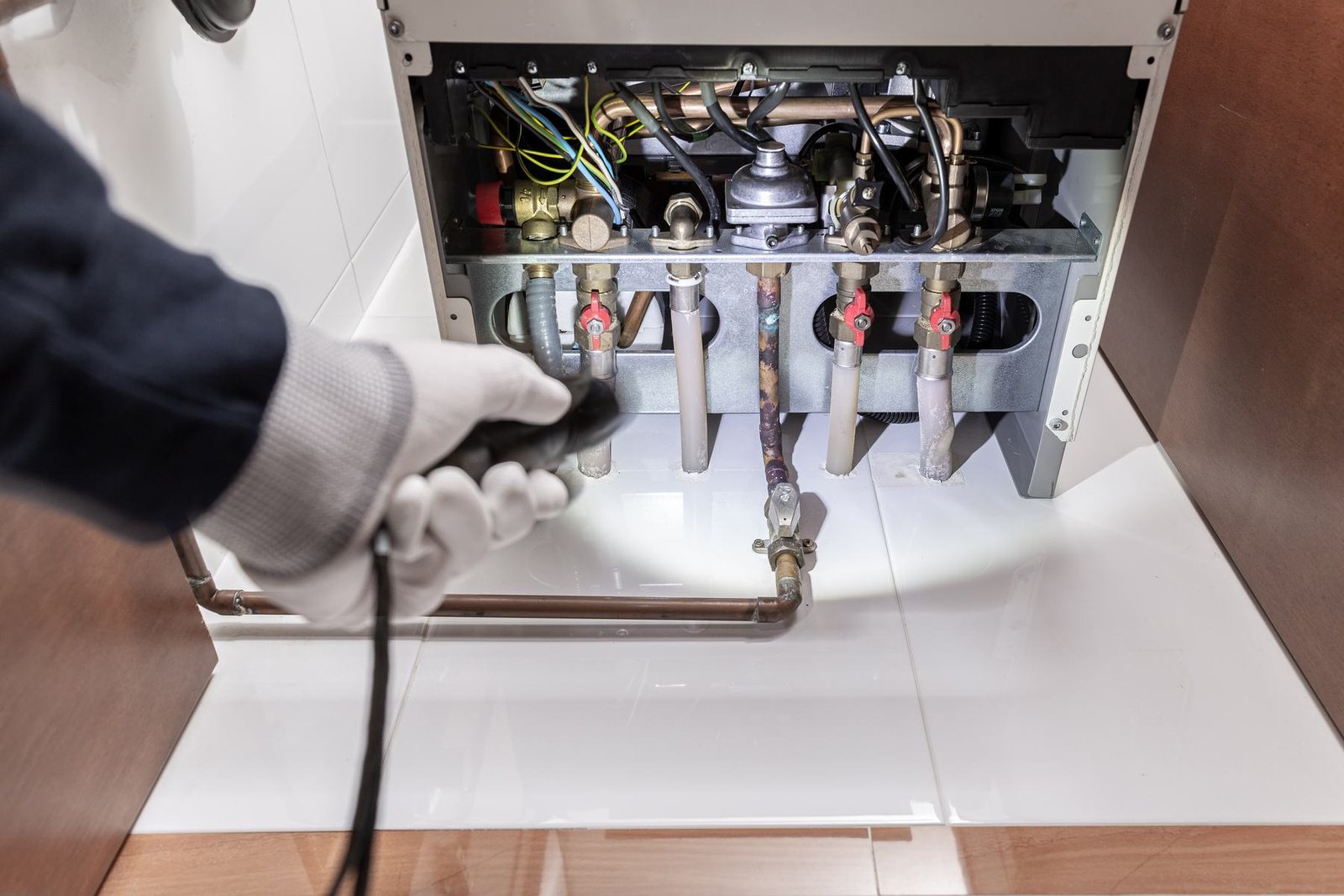 A technician in protective gloves servicing the internal valves and pipework of a domestic geyser.