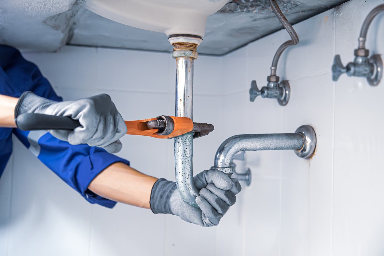 A plumber in work gloves tightening the trap under a bathroom basin with a pipe wrench.